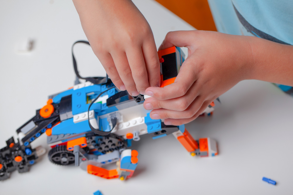 圖片展示一雙兒童的手正在組裝一個用 LEGO 積木構建的機械人，組件以藍、白、橙為主色調，擺放在白色桌面上。場景充滿創意與學習氛圍，突顯 STEM 教育中動手實作的重要性。